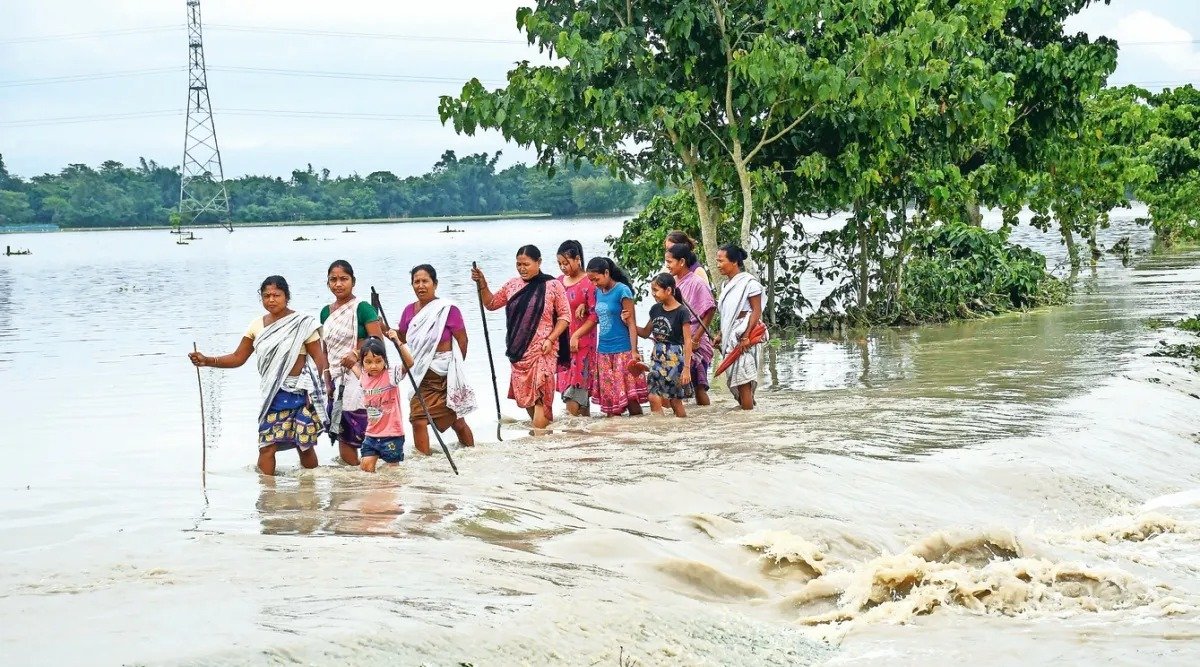 Women and a young girl walking through waist-deep floodwater with bamboo sticks in Assam